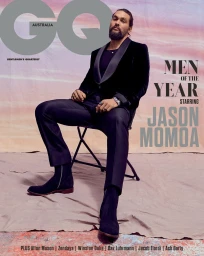 Jason Momoa seated on a folding chair wearing a dark velvet blazer and black trousers against a painted backdrop.