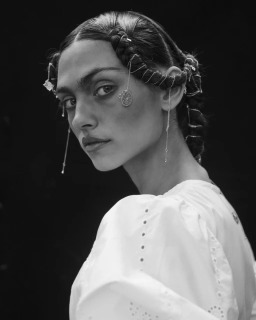 Portrait of a fair-skinned woman with styled braids, silver chains in her hair, and a white cotton top, set against a dark background.
