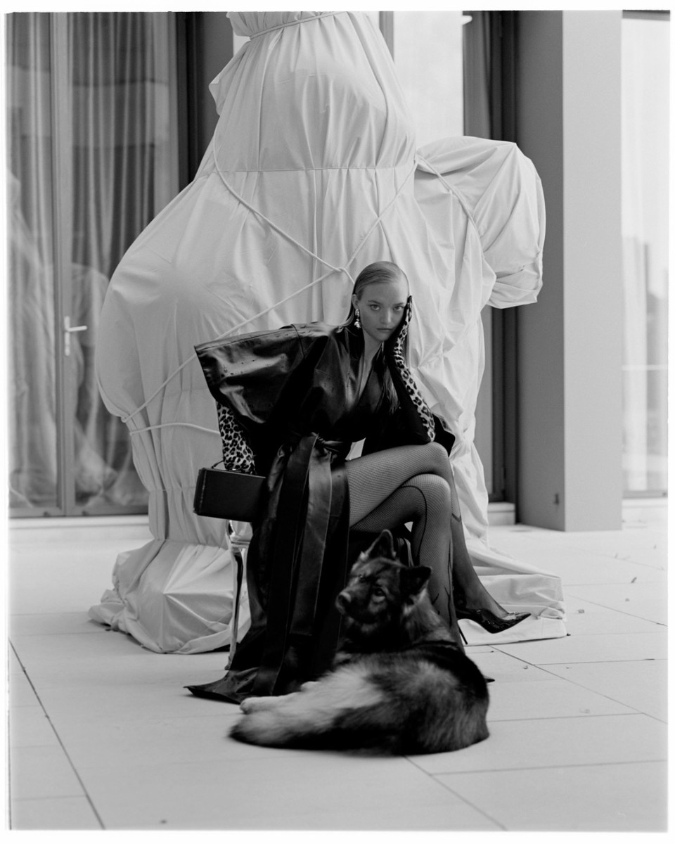 Black and white portrait of a fair-skinned adult female model in a dramatic black coat, seated with a dog, alongside an object wrapped in fabric indoors.