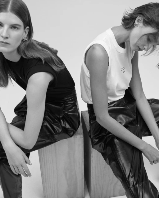 Two women in fashionable black pants and shirts sit on wooden blocks in a studio. Black and white fashion shot with minimalist background.