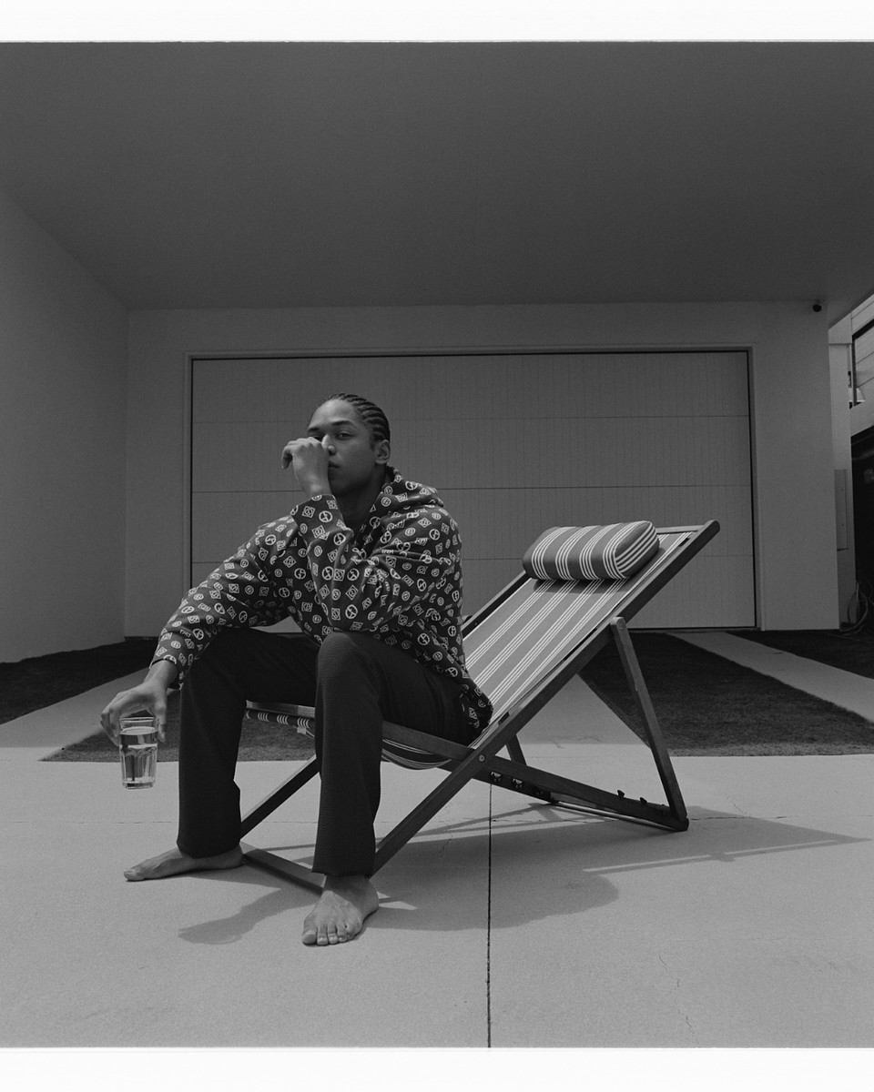 Black and white shot of a man seated on a beach chair, holding a glass. He wears patterned top, trousers, with a garage in the background.