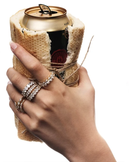A person holds a beer can wrapped in a baguette, adorned with several diamond rings on their fingers against a white backdrop.