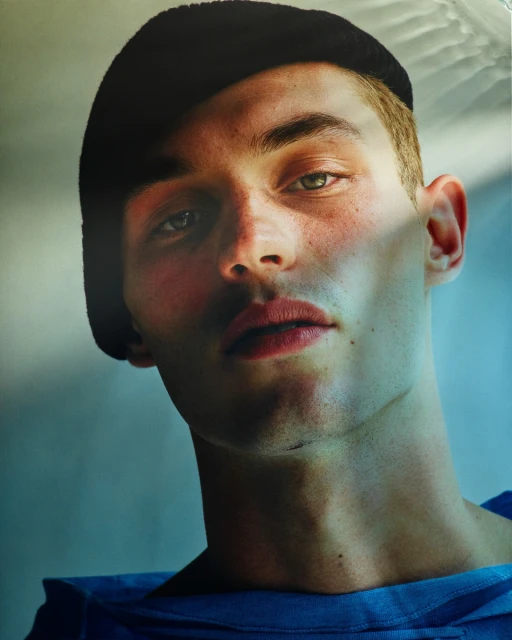 Close-up portrait of a young adult man with short hair, wearing a dark cap and blue top, showcasing cool lighting and natural skin texture.