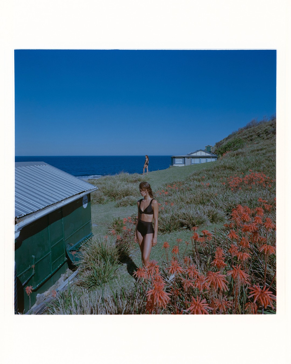 Woman in a black bikini standing on a grassy coastal slope near red flowering plants and a green building by the sea