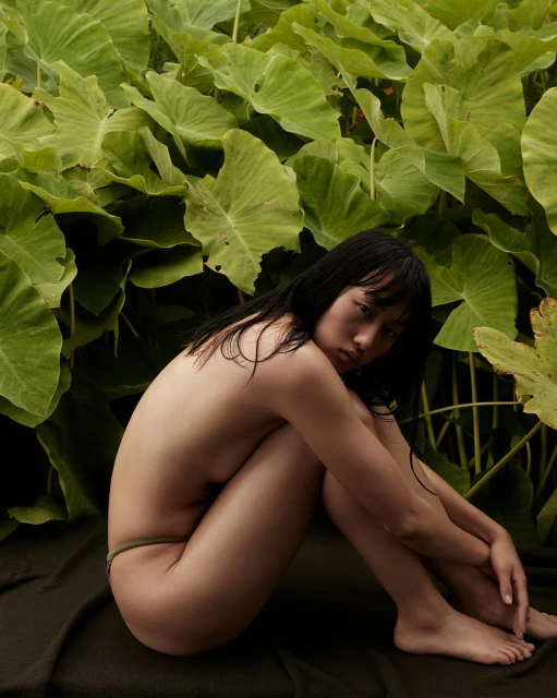 Seated nude Asian woman poses surrounded by lush green foliage, creating a natural, intimate portrait in a serene outdoor setting.