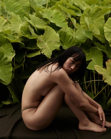 Seated nude Asian woman poses surrounded by lush green foliage, creating a natural, intimate portrait in a serene outdoor setting.