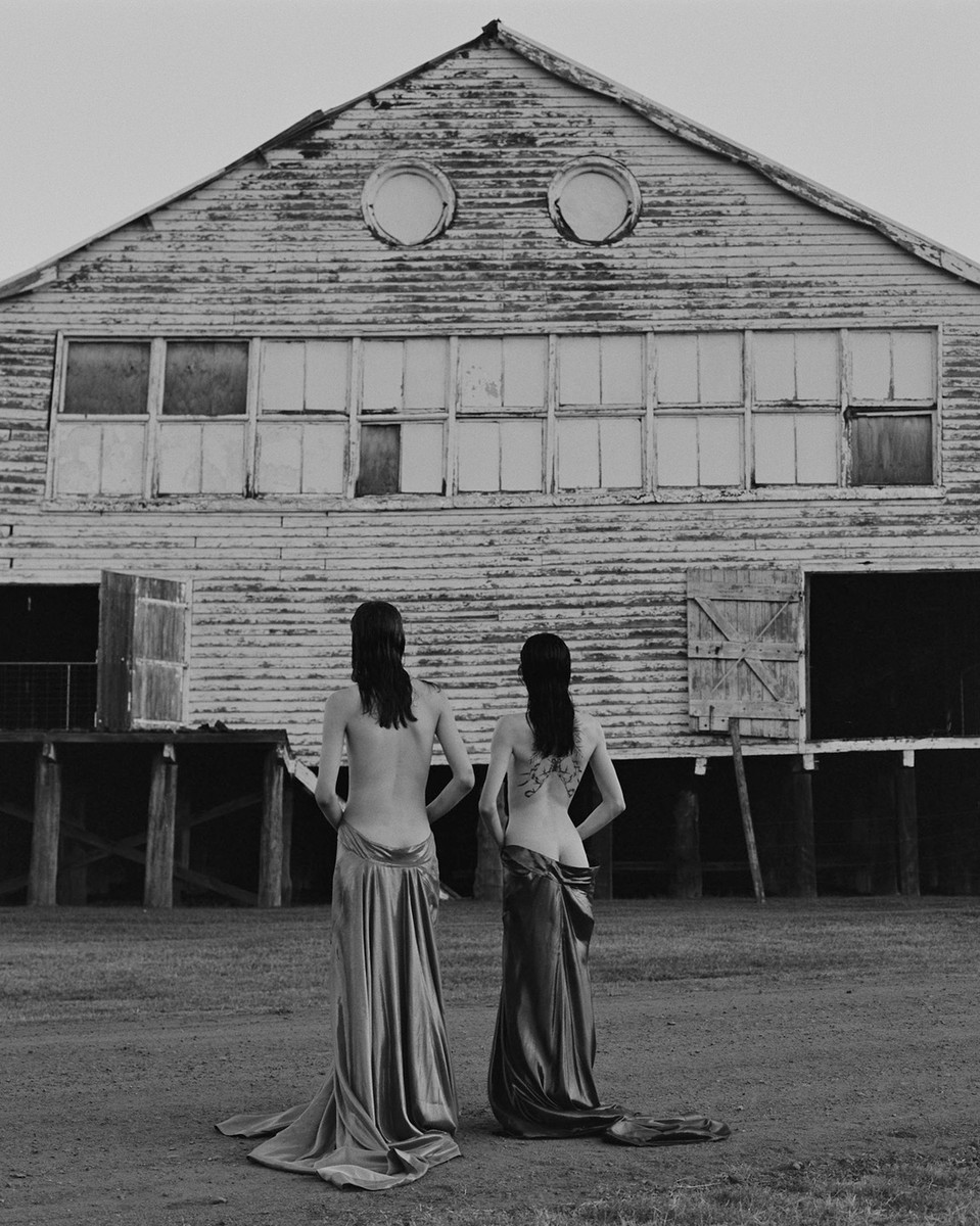 Two people standing with backs to camera wearing draped skirts in front of weathered wooden building exterior