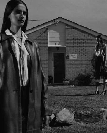 Two women pose outdoors in black and white, wearing long coats, with a brick house and lawn in the background.