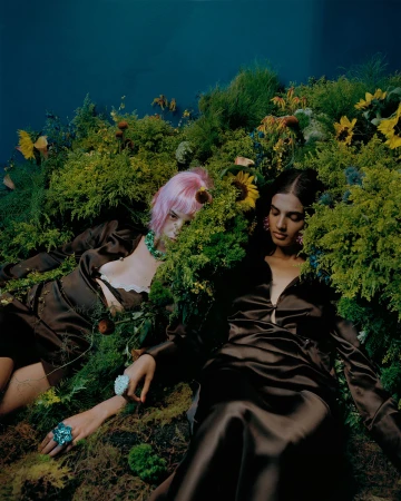 Two women in satin dresses lie amidst lush greenery and wildflowers, creating a harmonious blend of fashion and nature.