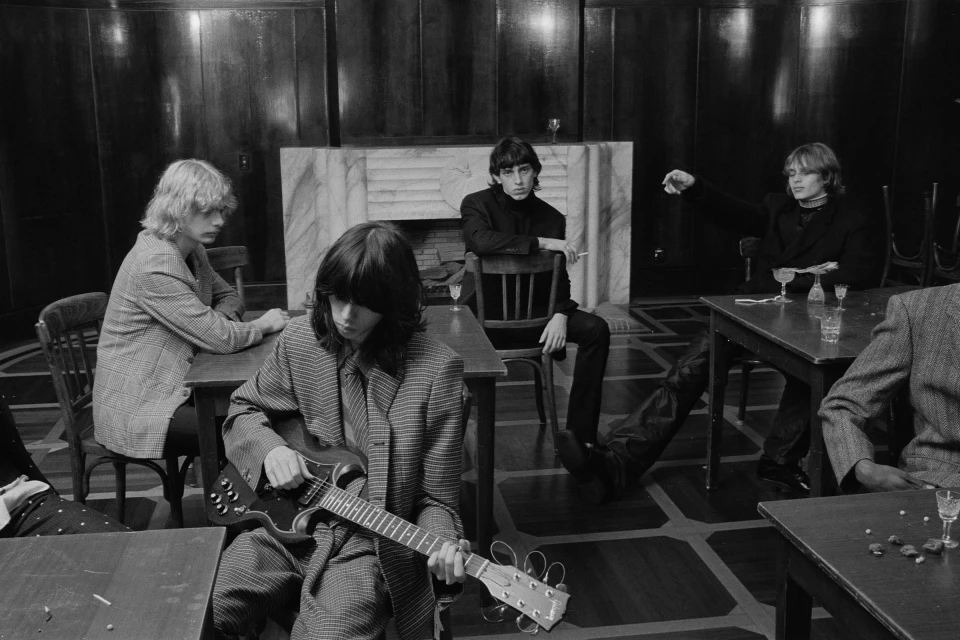 Five people sit at tables indoors; one plays electric guitar. The group is dressed in blazers. The scene is shot in black and white.