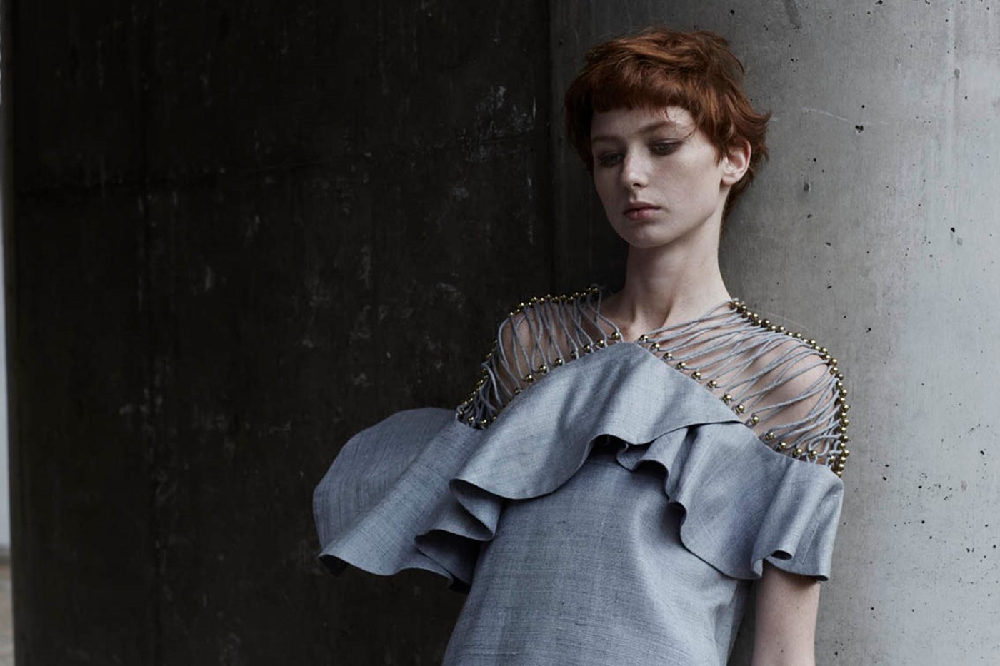 Fashion shot of a woman with short red hair posing in a light gray ruffled dress with beaded straps against a dark concrete background.