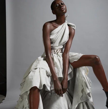 Model in a draped one-shoulder white striped dress with a belted waist against a neutral grey seamless studio background.