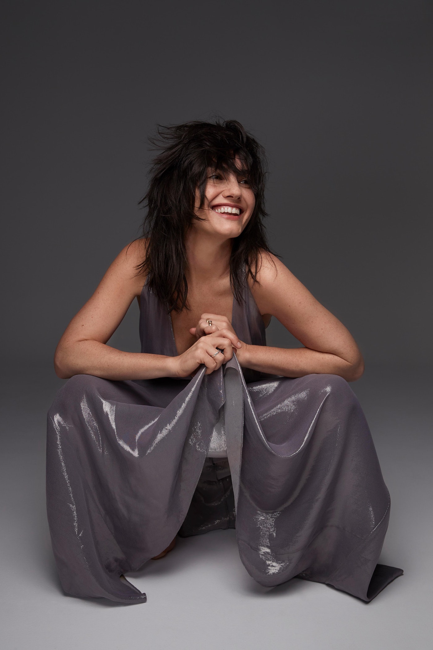 Smiling woman in a gray jumpsuit sitting on the floor in a studio setting, posing and looking off to the side with a happy expression.
