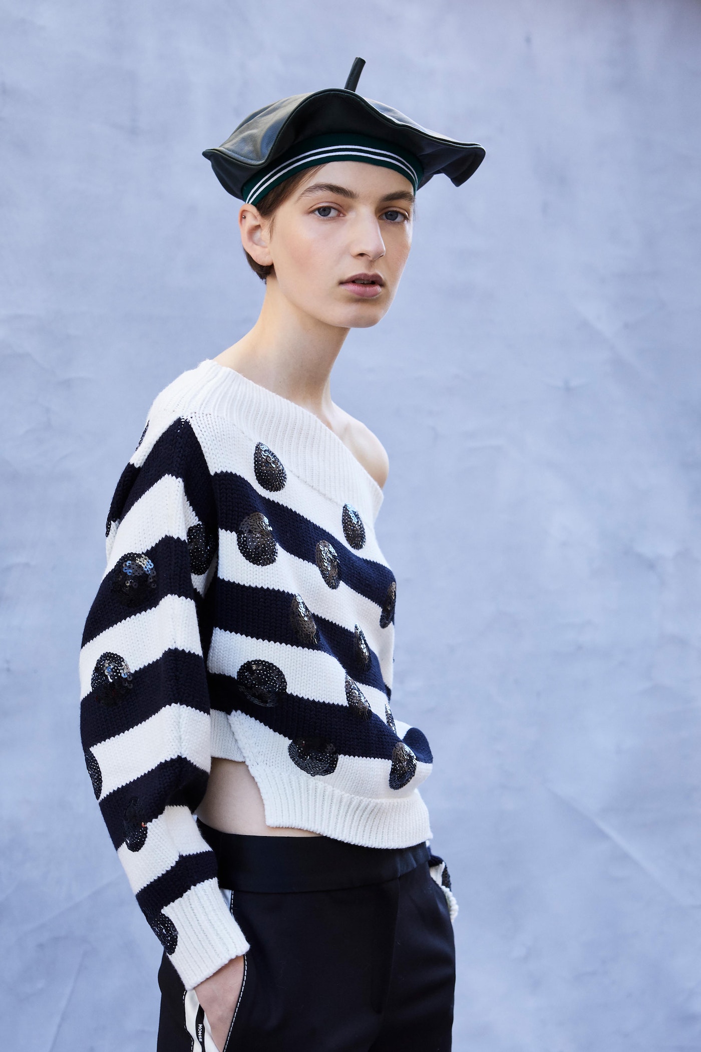 Woman wearing a black beret, and a striped off the shoulder sweater with polka dots in front of a blurred blue background.