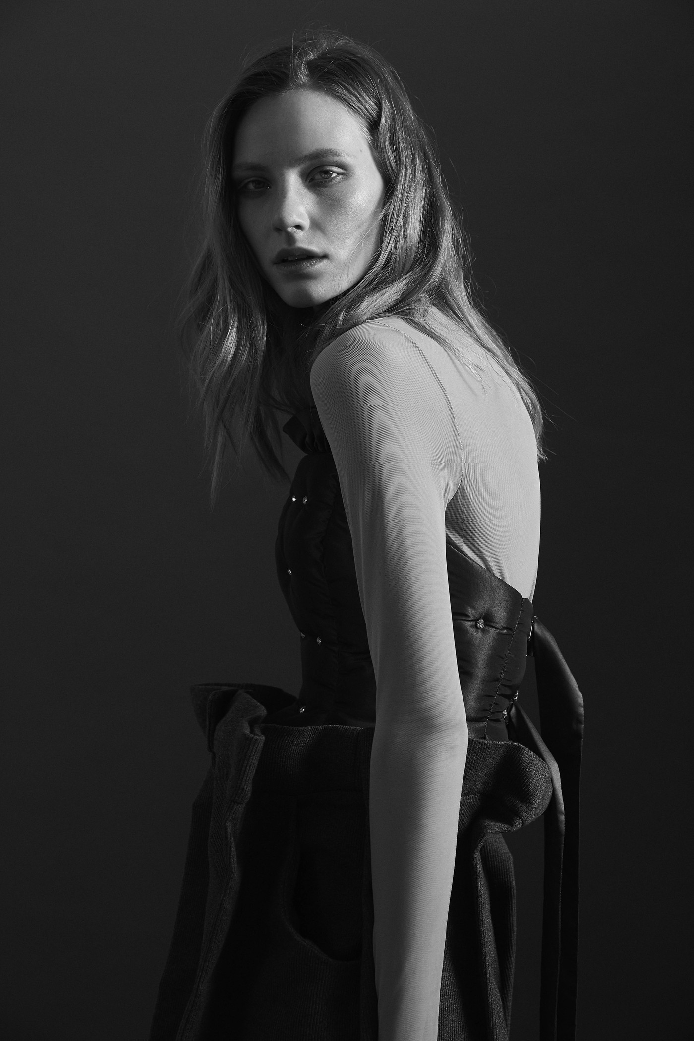 A striking black and white fashion portrait of a slender adult woman with long hair, wearing a dark, textured dress against a grey backdrop.
