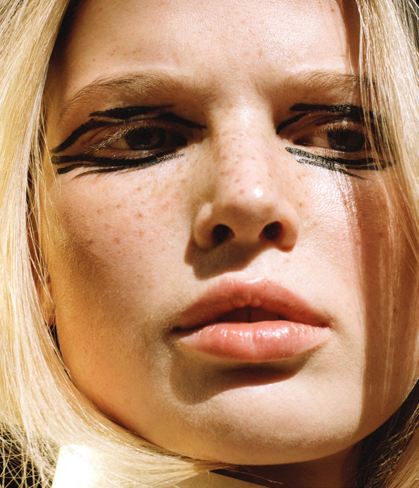 Close-up beauty shot of a woman with blond hair and graphic black eyeliner. The focus is on her face and makeup in soft lighting.