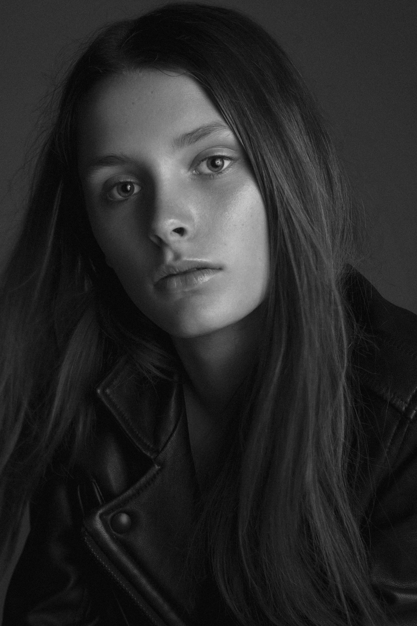 Monochrome close-up of a young adult female wearing a leather jacket, looking directly at the camera with long hair. Fashion shot.