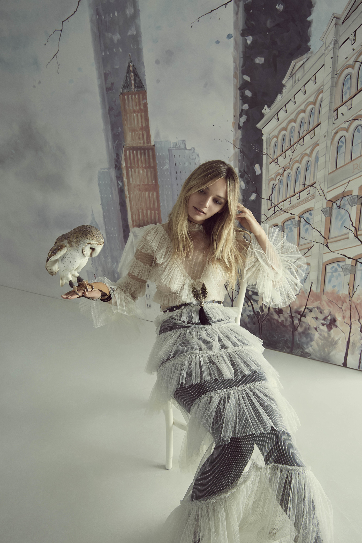 A blonde woman in a frilly white dress poses with an owl perched on her hand, set against a painted backdrop of city buildings.