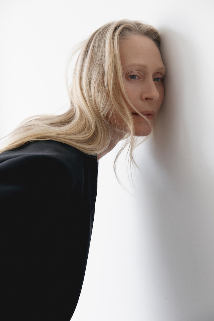 Adult woman with blonde hair, wearing a black blazer, leans her face against a white wall in a studio setting, looking pensive.