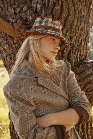 A blond woman wearing a plaid hat and matching plaid blazer leaning against a tree trunk outdoors in Sydney.