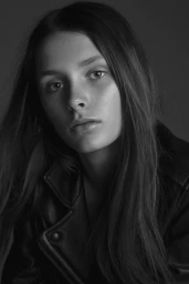 Close-up monochrome portrait of a young woman with long dark hair wearing a black leather jacket against a plain dark background.