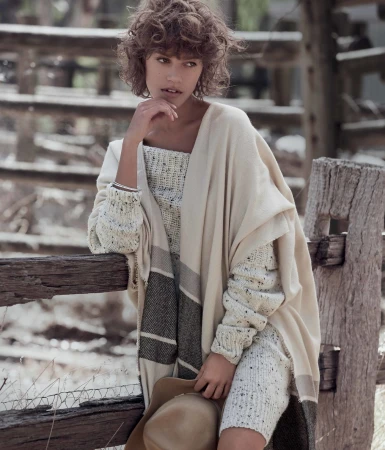Woman with curly brown hair leaning on a weathered wooden fence wearing a cream sweater and poncho