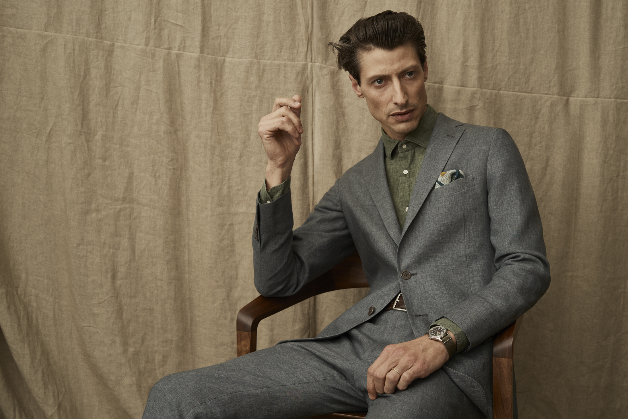A confident adult man in a grey suit is sitting on a chair in an indoor setting, posing for a fashion or lifestyle shot, wearing a watch.