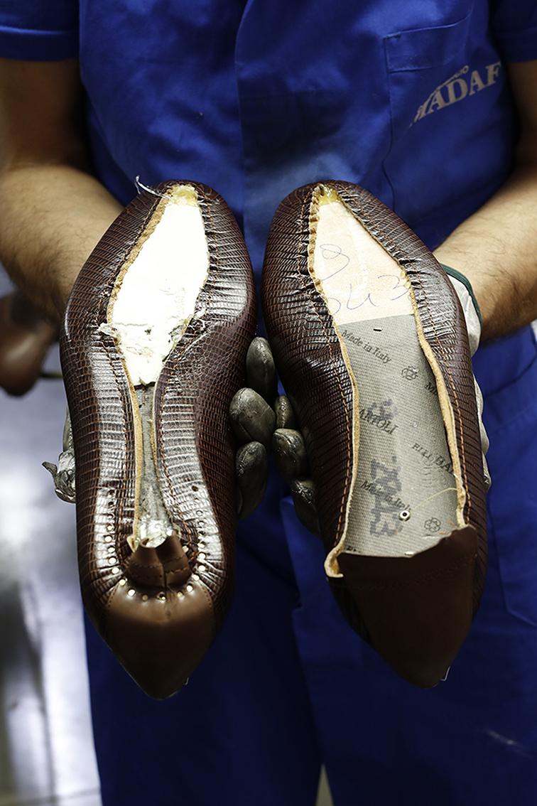 Close-up of gloved hands holding inside-out brown lizard-skin high heels, revealing their construction in a manufacturing setting.