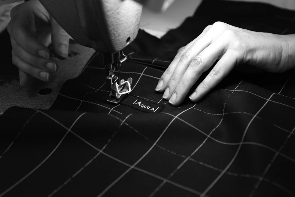 Close-up shot of a sewing machine and hands working on a black and white checkered fabric with the brand name Aquila visible.