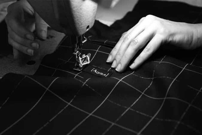 Close-up shot of a sewing machine and hands working on a black and white checkered fabric with the brand name Aquila visible.