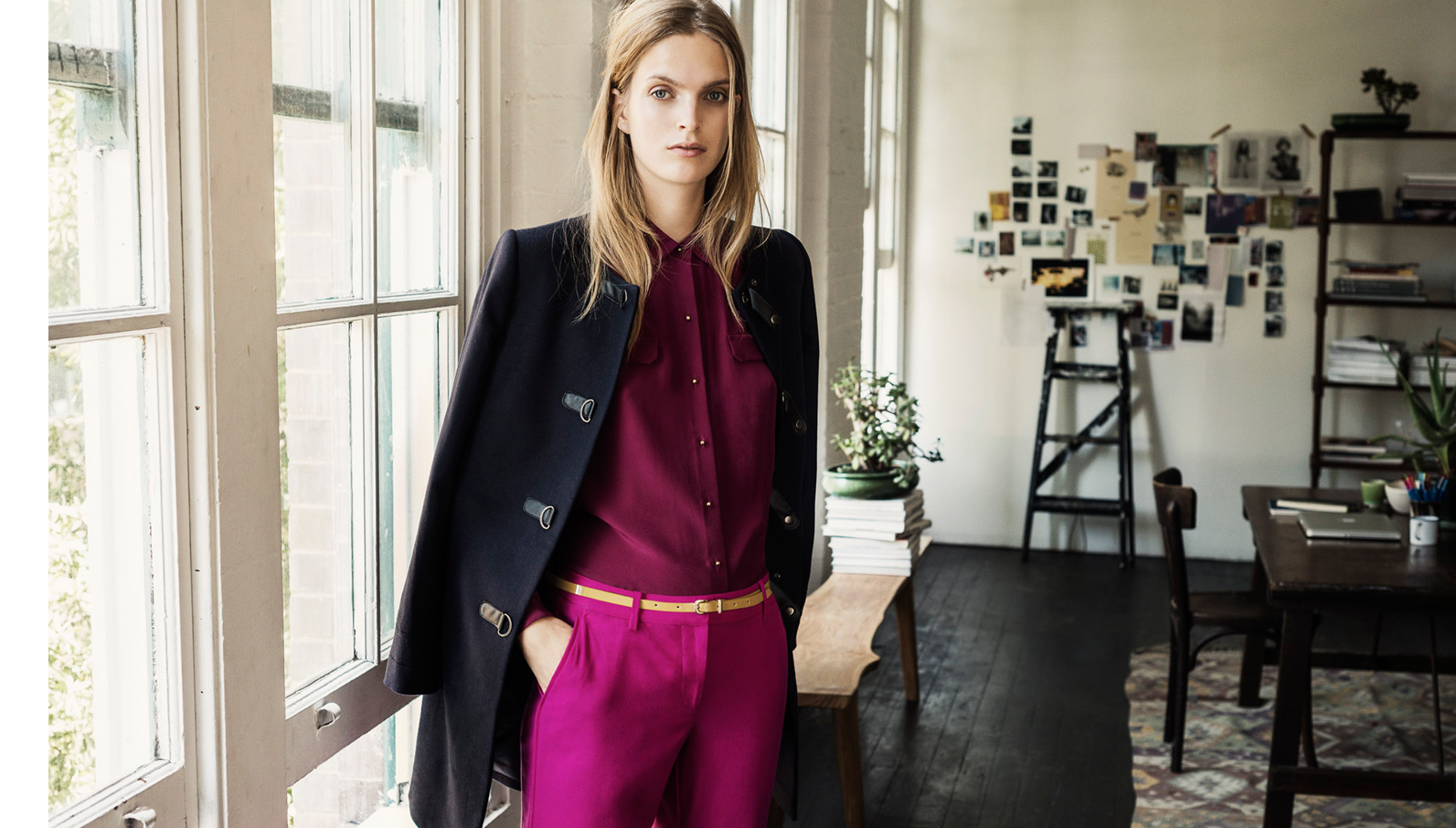 Blonde woman in magenta pants and button-down, with a navy coat draped over shoulder, poses near a window in an indoor setting.