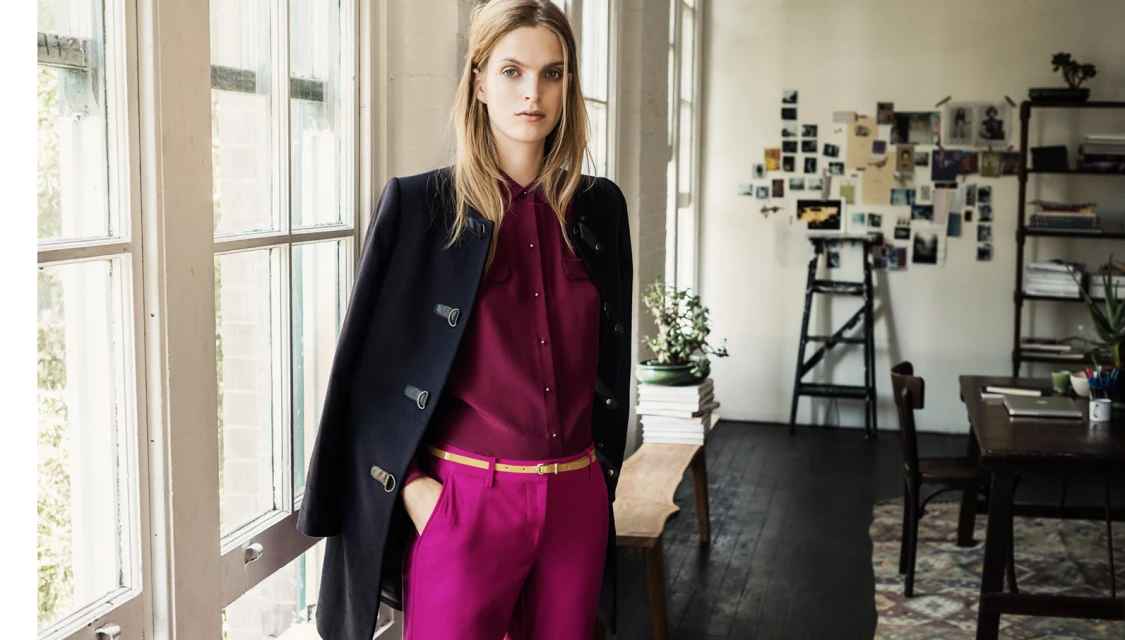 Blonde woman in magenta pants and button-down, with a navy coat draped over shoulder, poses near a window in an indoor setting.