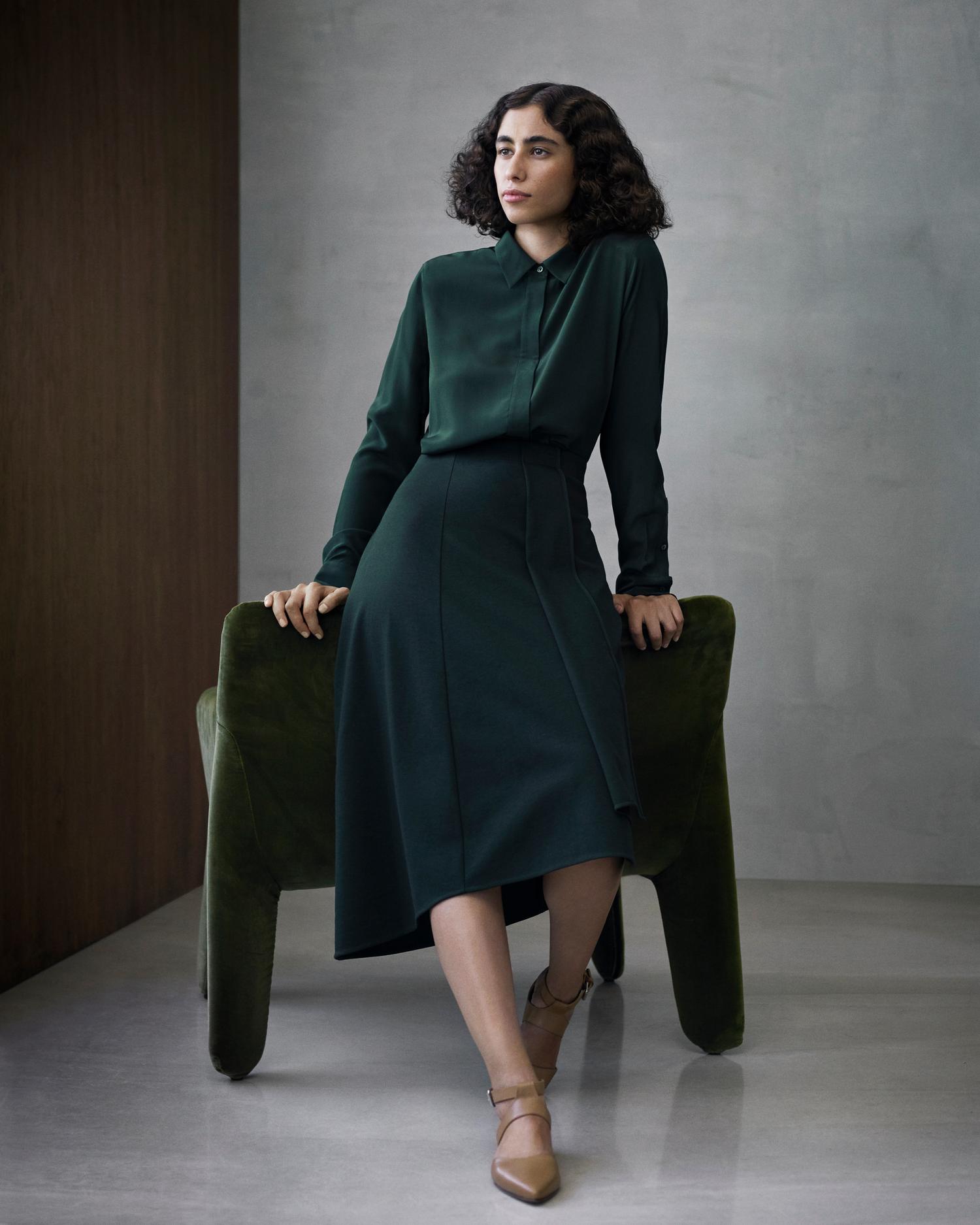 Woman in dark green long sleeve blouse and midi skirt sitting on green velvet chair in a studio setting