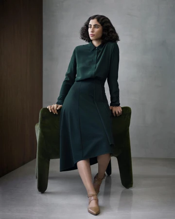 Woman in dark green long sleeve blouse and midi skirt sitting on green velvet chair in a studio setting