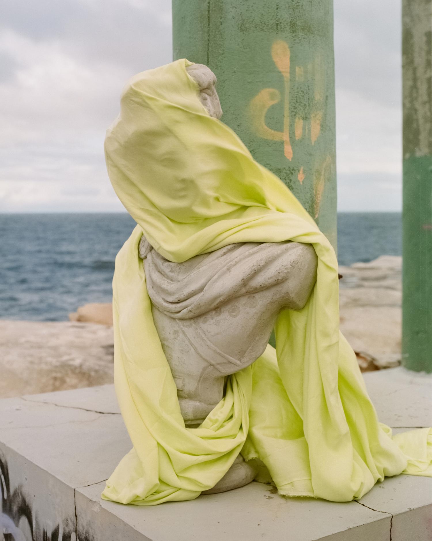 Statue of a seated figure draped in yellow fabric on a concrete pier near the ocean under a cloudy sky. Light, airy and serene.