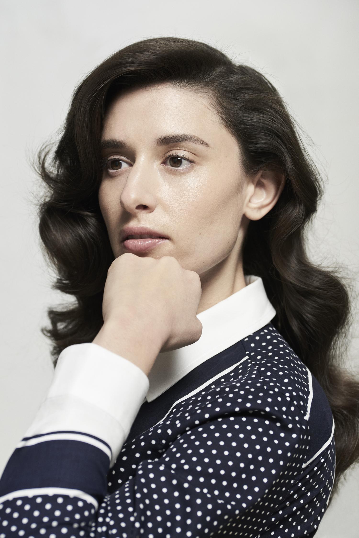 Headshot of an adult female with wavy dark hair, wearing a navy polka dot shirt with a white collar, posing in a bright studio setting.