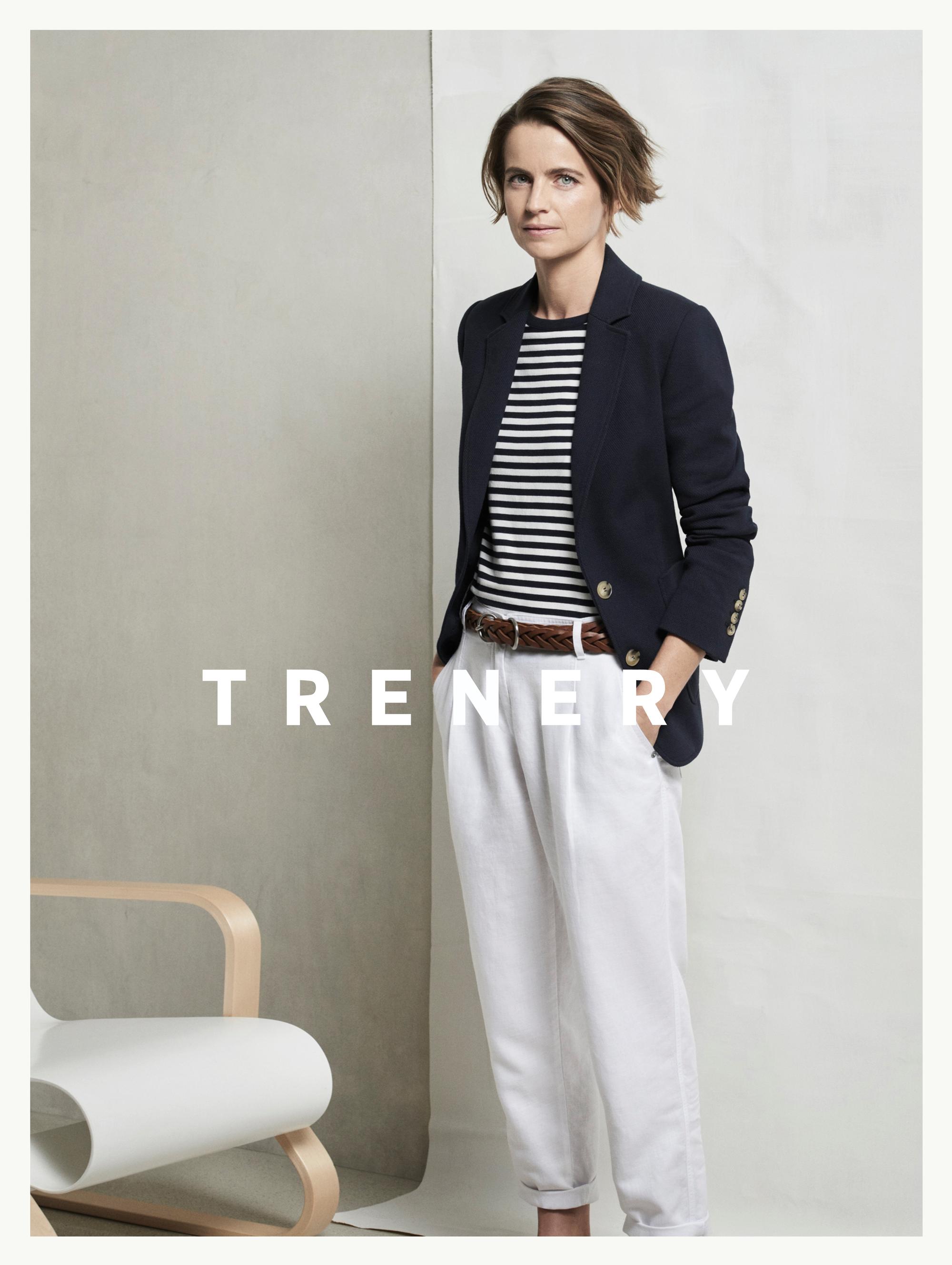 A woman in a blazer and striped shirt poses indoors next to a modern chair, modeling a fashion-forward, minimalist style for Trenery.