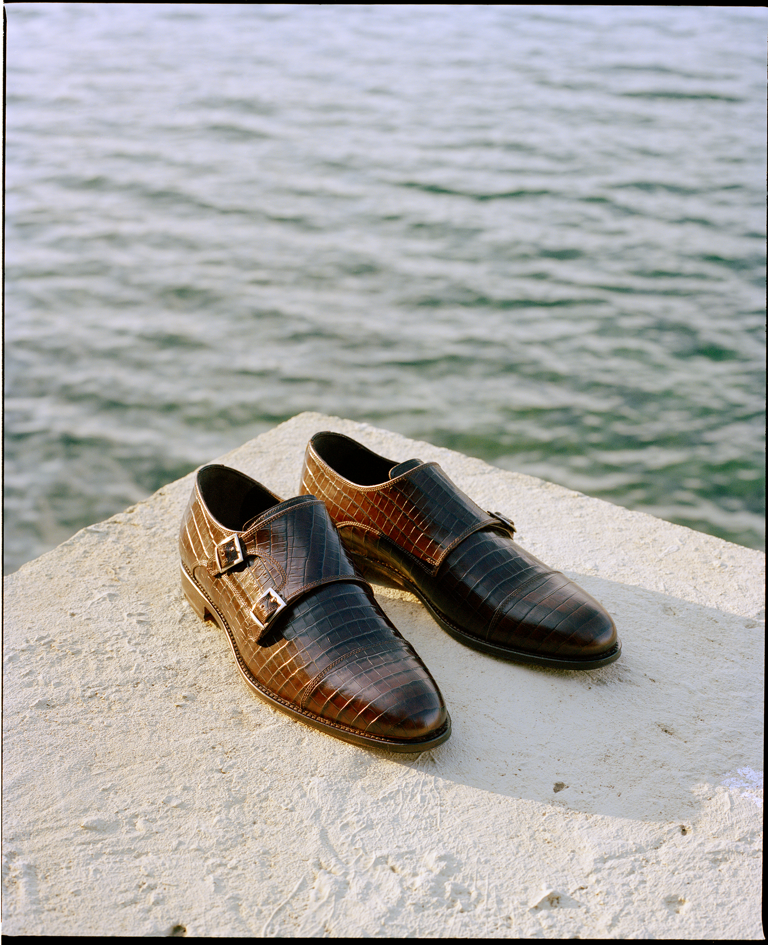 A pair of brown leather double monk strap shoes with an alligator skin pattern sits on a white concrete surface with a sea background.