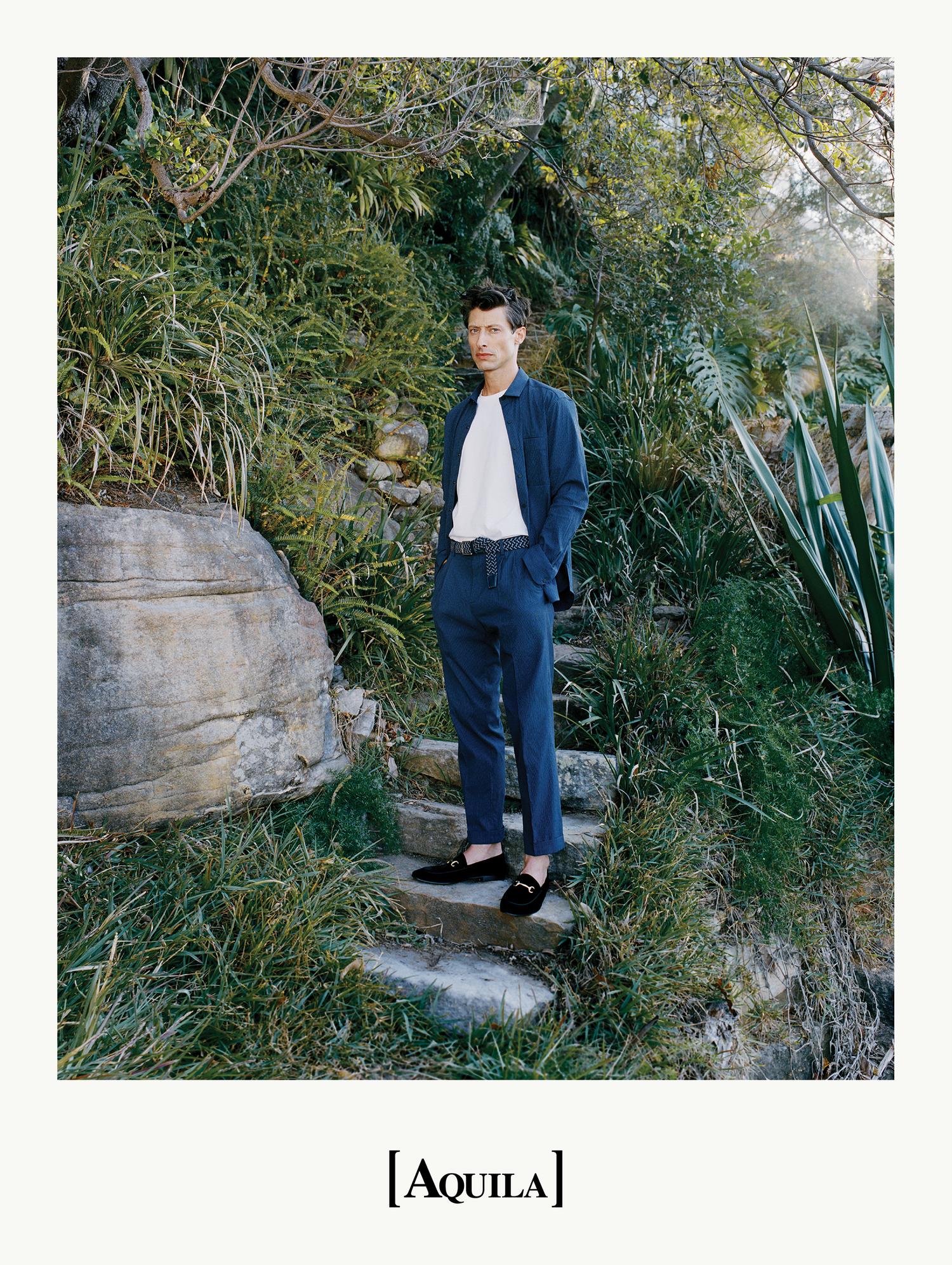A stylish adult male stands on stone steps in a lush garden, wearing a navy suit and loafers, against a backdrop of greenery.