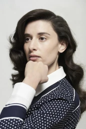 Portrait of a woman with dark wavy hair wearing a navy blue polka dot shirt with a white collar, resting her chin on her hand.