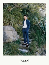 Man wearing a blue button-down shirt and trousers standing on stone steps surrounded by lush foliage with visible text Aquila at the bottom.