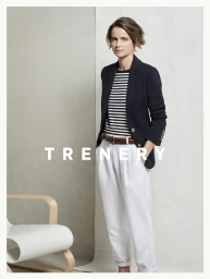 Woman wearing a dark blazer, striped top, and white trousers standing against a split grey and white studio background.