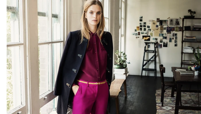 Woman standing indoors near large windows wearing a dark coat, purple blouse, and pink trousers.
