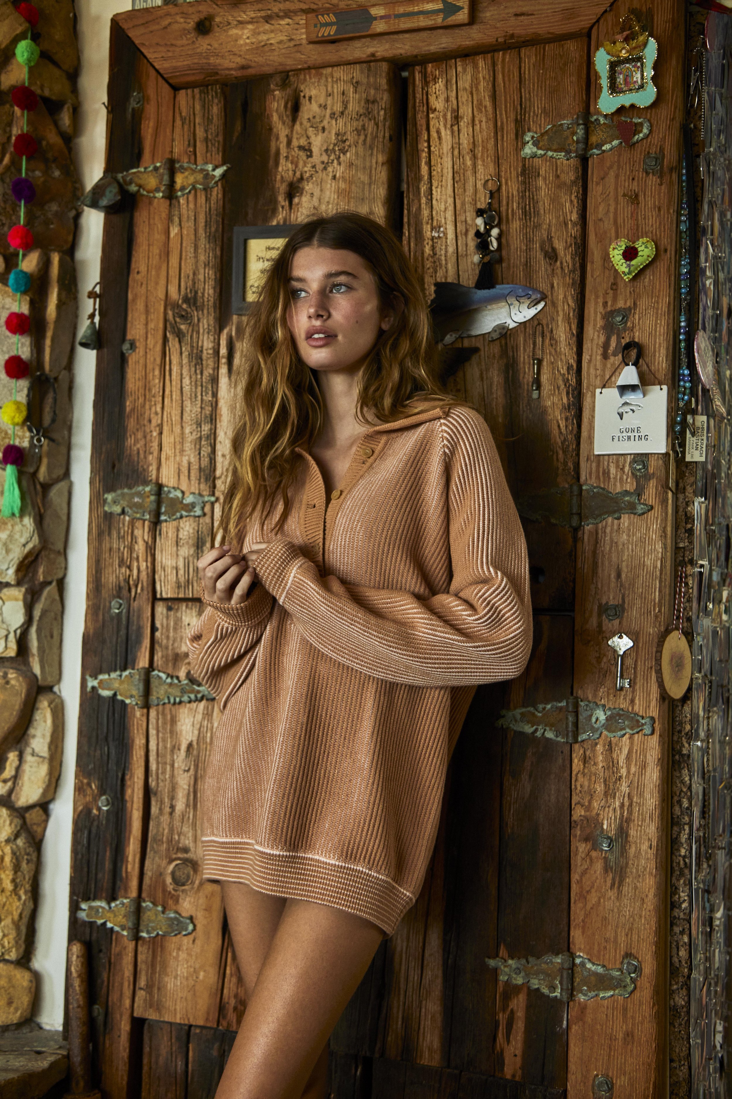 A woman with wavy blonde hair is posing indoors wearing a tan collared sweater, in front of a rustic wood door with decorative elements.