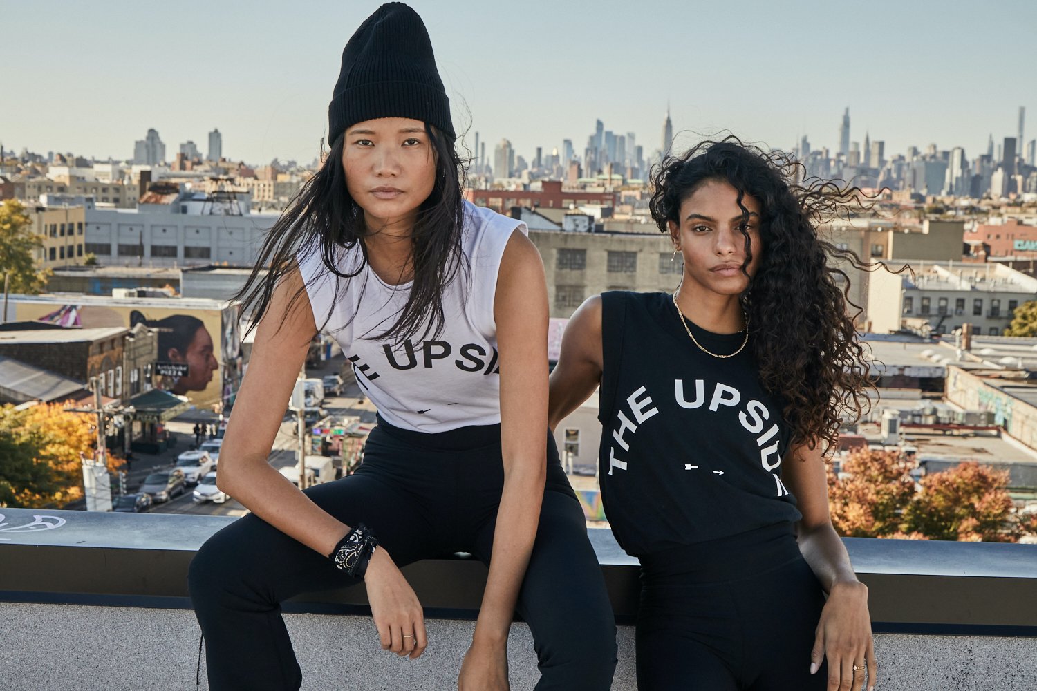 Two athletic women in sleeveless tops and leggings pose on a rooftop with an urban skyline backdrop under daylight.