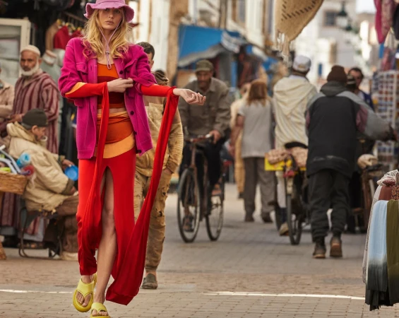 Fashionable model in a pink jacket, red dress, and yellow sandals walks down a street in an outdoor market with people and bicycles.