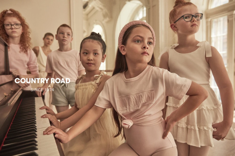 Group of children in light pink clothing stand and sit near a piano, looking forward with neutral expressions in a well-lit room.