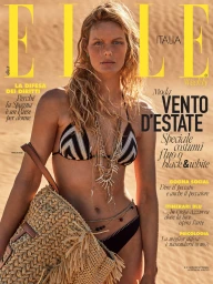 A female model wearing a black and white patterned bikini and layered necklaces holding a straw beach bag in front of a sandy background.