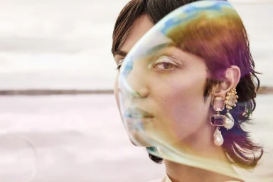 Close-up portrait of a woman wearing ornate gemstone and pearl drop earrings behind a translucent iridescent bubble.