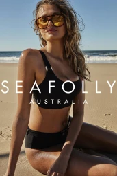 Model wearing oversized tinted sunglasses and a black bikini seated on a sunny sandy beach by the ocean.